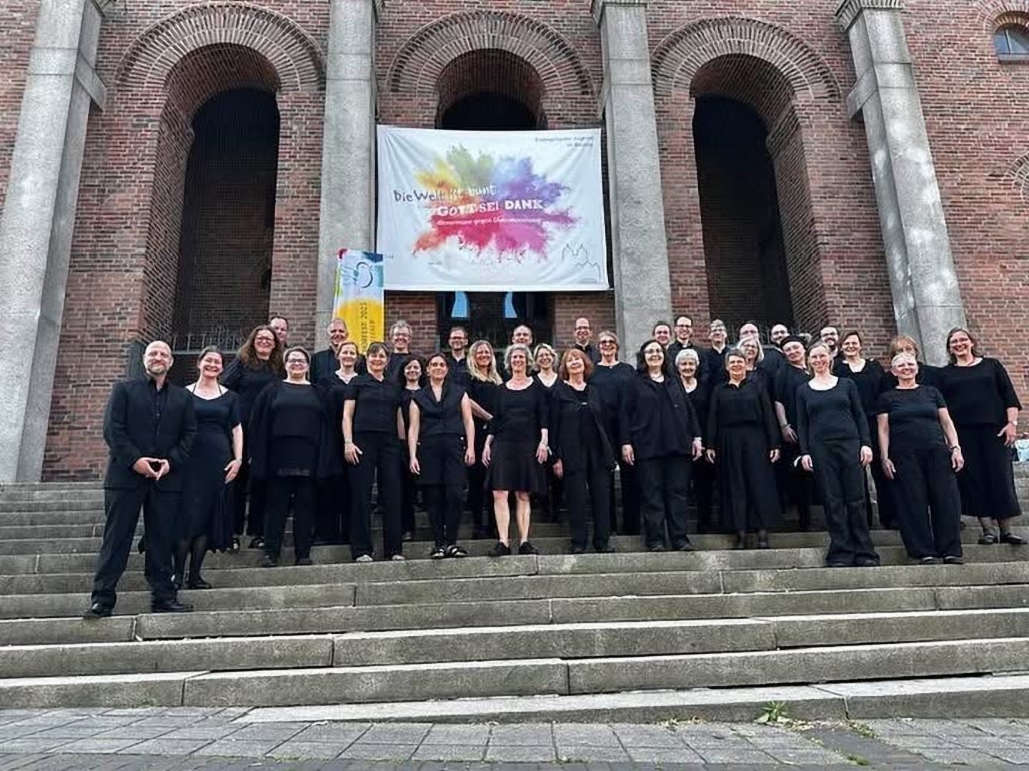 Die Capella Vocalis vor der Kirche mit der Fahne „Die Welt ist bunt – Gott sei Dank“